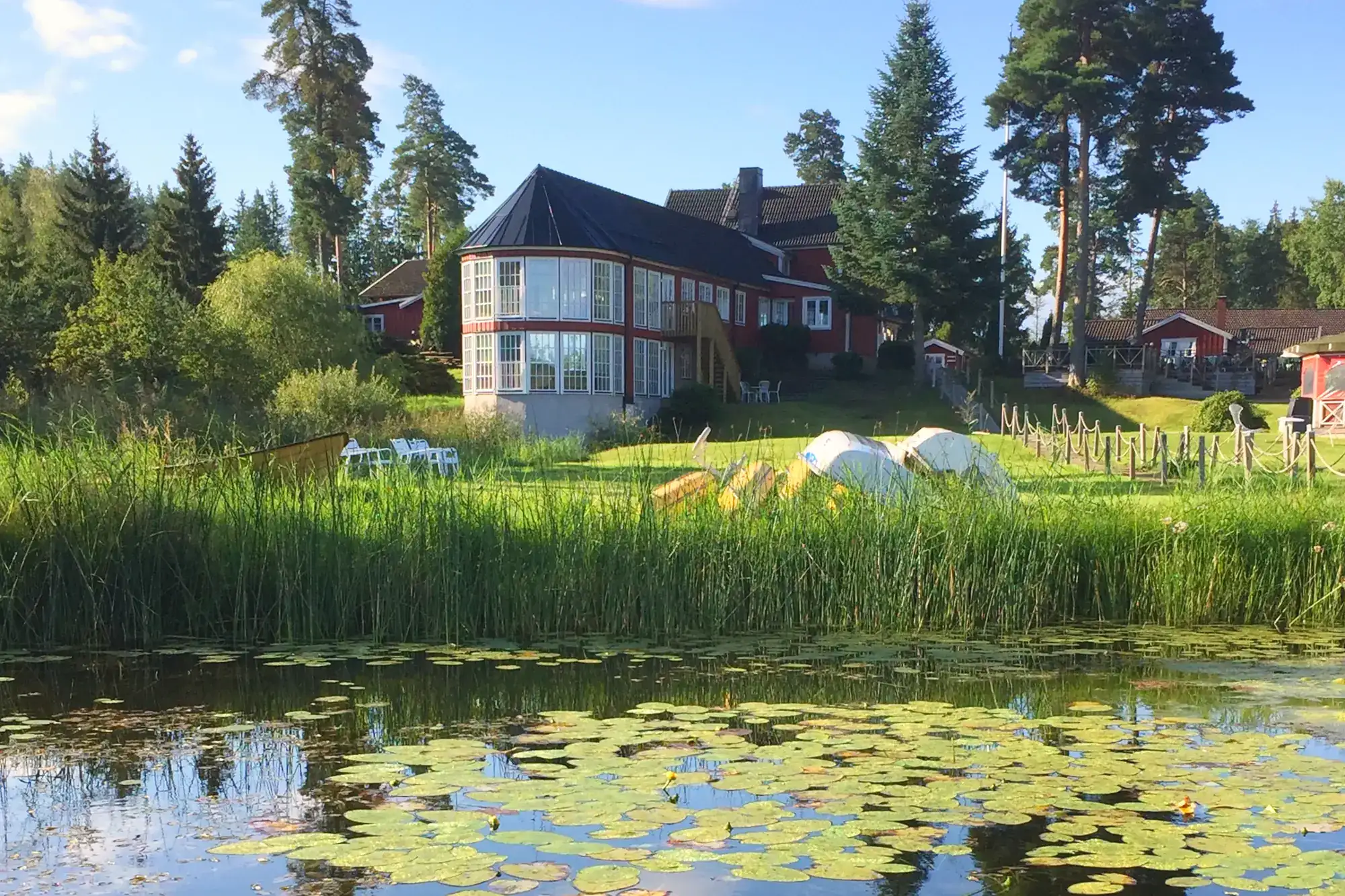 Odalgårdens röda huvudbyggnad med glasat burspråk, sedd över sjön med näckrosor och vass en solig sommardag.