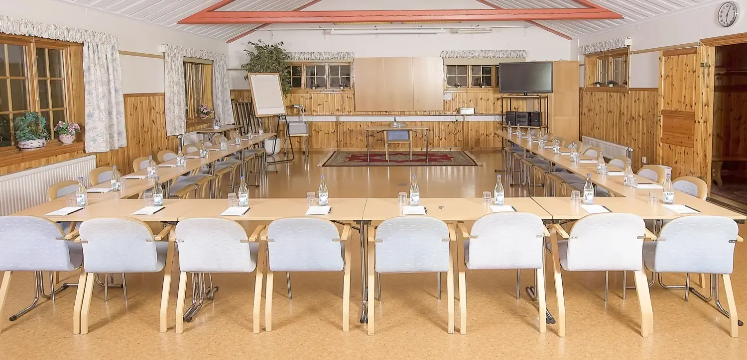 Konferenssal med bord i U-form, ljusa stolar och fönster längs väggarna.
