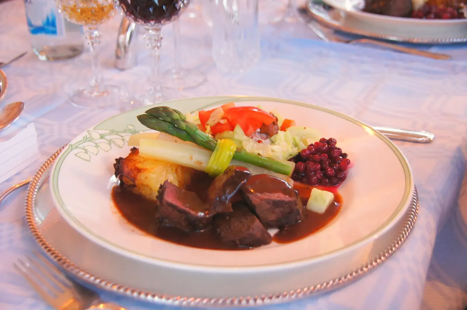 Varmrätt med kött, sparris, grönsaker och lingon på en vit tallrik vid ett dukat bord.