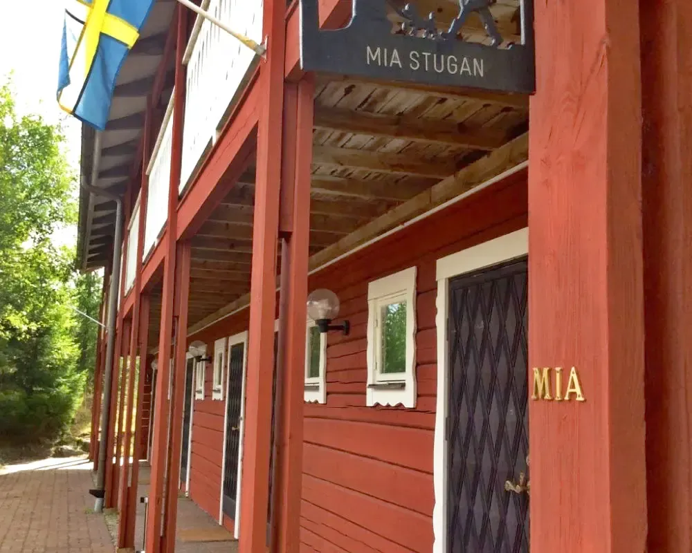 Röd stugbyggnad med vita fönsterfoder och två svenska flaggor vid entrén.